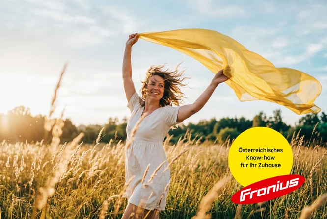 Eine junge Frau in einem weißen Kleid tanzt ausgelassen auf einer sonnenbeschienenen Wiese und hält einen wehenden gelben Schal über den Kopf. Die Szene spielt sich in hohem, goldenem Gras unter einem warmen Nachmittagshimmel ab und vermittelt eine unbeschwerte Sommerstimmung. In der unteren rechten Ecke befindet sich ein gelber Kreis mit dem deutschen Text „Maximal Regional: Speicher Made in Austria“ neben dem roten Fronius-Logo. Das Gesamtbild strahlt Freiheit, Natur und regionale österreichische Qualität aus.