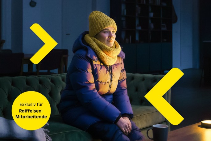 Eine Person mit gelber Mütze, gelbem Schal und blauem Wintermantel sitzt auf einem grünen Sofa in einem schwach beleuchteten Zimmer. Ein Holztisch mit einer Tasse und einer sanft leuchtenden Lampe trägt zur behaglichen Atmosphäre bei. Die Szenerie lädt zum Entspannen in der kalten Jahreszeit ein.