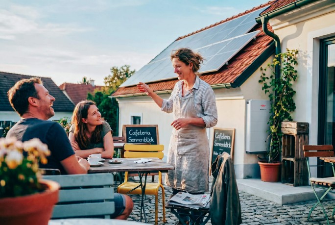 Café-Besitzerin unterhält sich mit Gästen im Schanigarten, im Hintergrund ist ein Stromspeicher sichtbar
