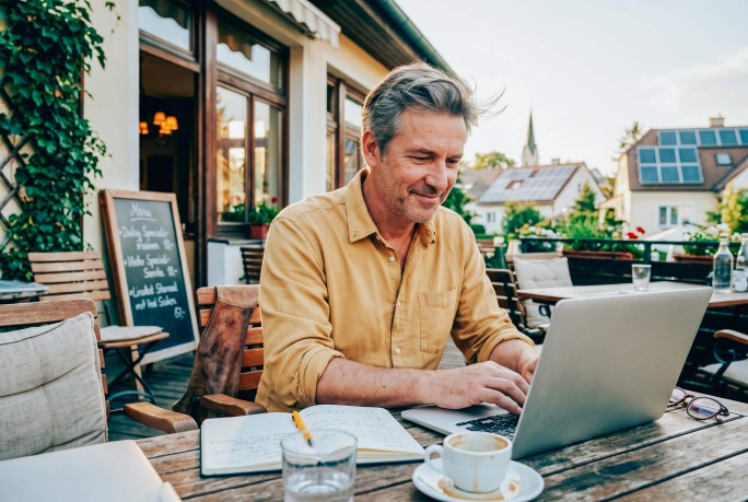 Lokalbesitzer sitzt am Tisch vor seinem Gasthaus und arbeitet am Laptop, daneben Kaffee und Notizbuch
