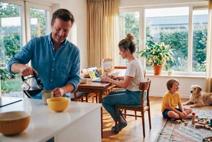 Familie beim Frühstück, Frau meldet sich am Laptop bei Raiffeisen Energie an, Mann schenkt Kaffee ein, daneben spielt das Kind
