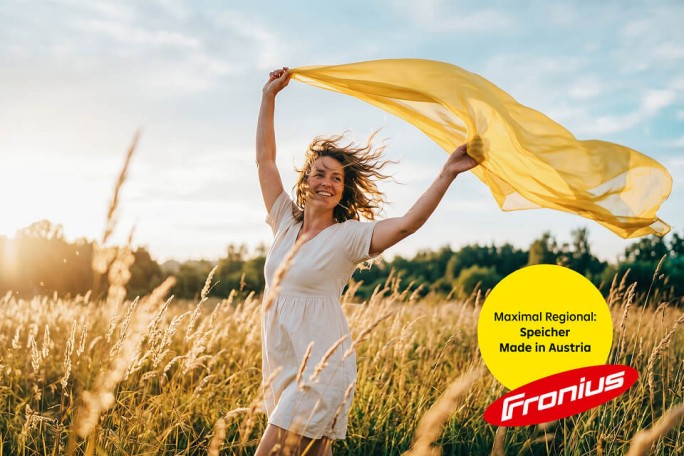 Eine junge Frau in einem weißen Kleid tanzt ausgelassen auf einer sonnenbeschienenen Wiese und hält einen wehenden gelben Schal über den Kopf. Die Szene spielt sich in hohem, goldenem Gras unter einem warmen Nachmittagshimmel ab und vermittelt eine unbeschwerte Sommerstimmung. In der unteren rechten Ecke befindet sich ein gelber Kreis mit dem deutschen Text „Maximal Regional: Speicher Made in Austria“ neben dem roten Fronius-Logo. Das Gesamtbild strahlt Freiheit, Natur und regionale österreichische Qualität aus.